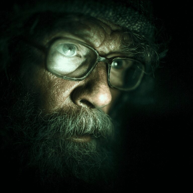 Elderly man with glasses in dim light.